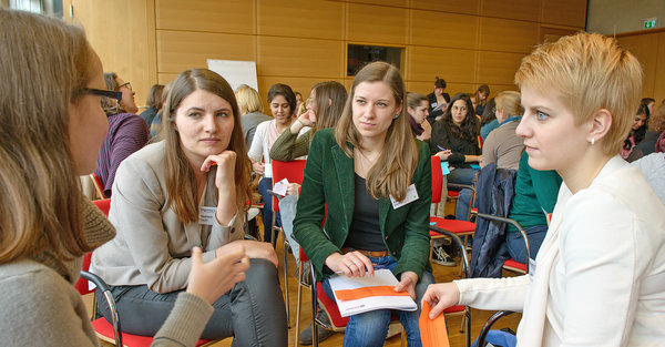 Full indoor shot of a group of young women at a conference or workshop. The women are seated in a circle, in red chairs, and are engaged in conversation. The setting is a room with light beige wooden paneling on the walls. The individuals are diverse in appearance, clothing styles and hair color, but all are dressed in casual business attire, such as jackets, blazers, and trousers/jeans. Several other women are seated in the surrounding area, participating in the group discussion.