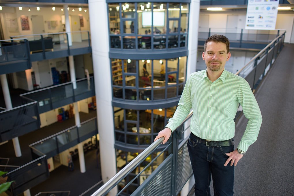 Christian Theobalt (wearing a light green shirt an jeans) is standig on an upper floor of the Max Planck Institute for Informatics which is open to inside. His hand rests on the steel banister of a rectangular gallery.