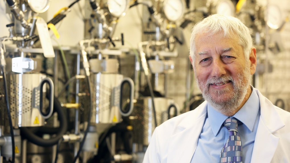 Portrait of Graham Hutchings wearing a lab coat over a blue shirt and blue patterned tie in front of cylindrical stainless steel reactors.