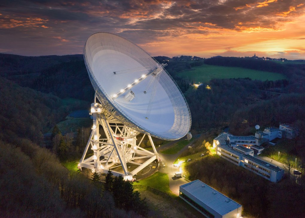 White parabolic antenna, turned half upwards, standing in a hilly and green landscape during sunset.