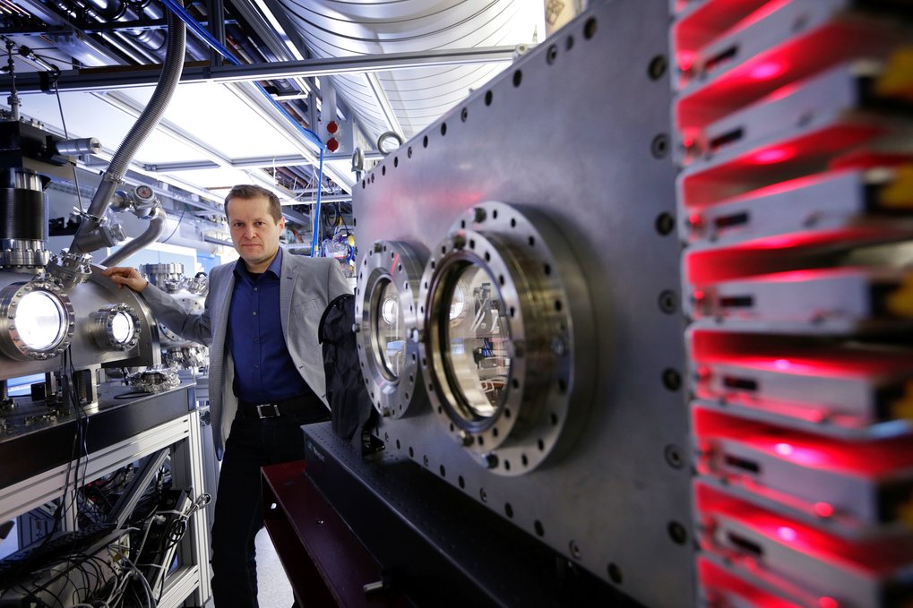 Ferenc Krausz is positioned diagonally behind a box-shaped vacuum chamber featuring two adjacent bull's-eye windows. A table-mounted camera occupies the right half of the picture. Krausz's right hand rests on another illuminated vacuum chamber, adorned with two portholes.