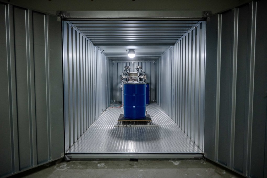 View into a brightly lit container made of corrugated iron, in the centre is a blue barrel surrounded by a construction.