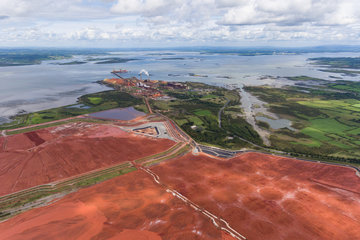 In the foreground, a section of the extensive area of a landfill site with rust-red mud, in the background a much smaller aluminium plant. The plant and landfill are located on a gulf, which can be seen in the upper half of the picture. Green meadows can be seen on the right.