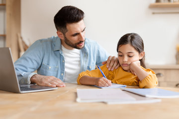 ein Vater mit seiner Tochter am Esstisch, auf dem Tisch steht ein Laptop, es liegen Blätter auf dem Tisch, das Mädchen schreibt etwas in ihr Heft. Die beidem machen zusammen Hausaufgaben.