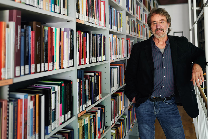 The picture shows researcher Steven Vertovec, Director at the Max Planck Institute for the Study of Religious and Ethnic Diversity, standing in front of a bookshelf in a library. Vertovec, wearing blue jeans, a blue shirt and a black jacket, is looking straight at the camera. He sports a full greying beard. 