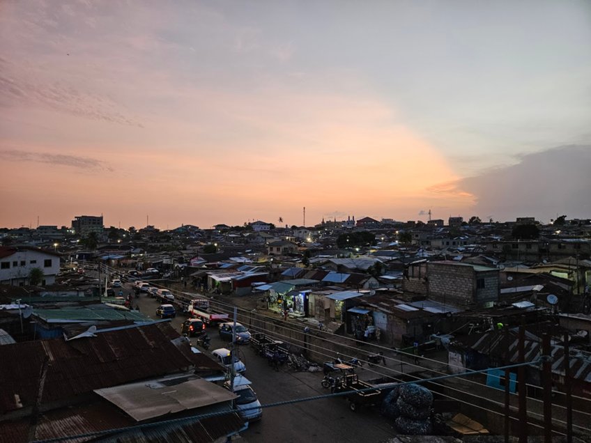 Picture of a street neighbourhood in Nima, a Zongo residential town in the region around Accra in Ghana. The picture is taken from above at what appears to be dusk, the light is diffuse and the sky tinged orange. You can see rooftops, and a street with houses of various sizes stacked against one another. Cars are driving down the street, some with their headlights on.