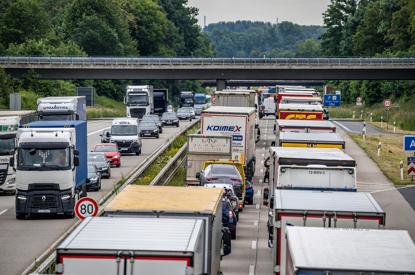 This picture, taken from a bridge over a motorway, shows heavy traffic with many lorries jammed on the two lanes in the direction of the camera. In the opposite direction, a few lorries and cars can be seen driving. A road bridge crosses the motorway a short distance away.