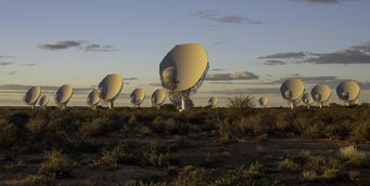 Numerous white parabolic dishes illuminated by the sunset stand on desert soil, looking in one direction.