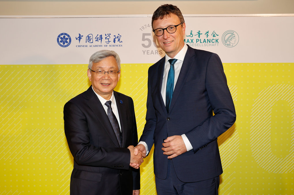 Two men in business suits are shaking hands. The man on the left, with short, graying hair, is wearing a dark navy or black suit, a light-colored collared shirt, and a dark tie. He is middle-aged, and appears to be of East Asian descent, judging by his facial features. He is smiling and looking directly at the camera. The man on the right, with short brown hair and glasses, is also wearing a dark navy or black suit, a light colored collared shirt, and a teal-blue tie.  He is middle-aged and appears to be of European descent. He is also smiling and looking directly ahead, though his focus is slightly turned toward the camera. The background is a bright, mustard-yellow backdrop with a repeating patterned design that resembles a grid or woven texture. Logos or banners, containing Chinese and English text, are visible on the wall behind the men. The banners seem to be related to science or an academic institution. The typography and graphic elements suggest a celebratory event or an anniversary.  The 1973-2023 or similar numbers and anniversary wording are on the backdrop.