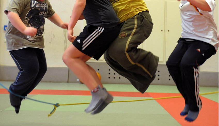 In this picture, you see four children in mid-jump in a gym environment. The children are wearing t-shirts and gym shorts or trouser. Only their legs and torsos are visible in the picture, and you can tell that the children are slightly overweight. 