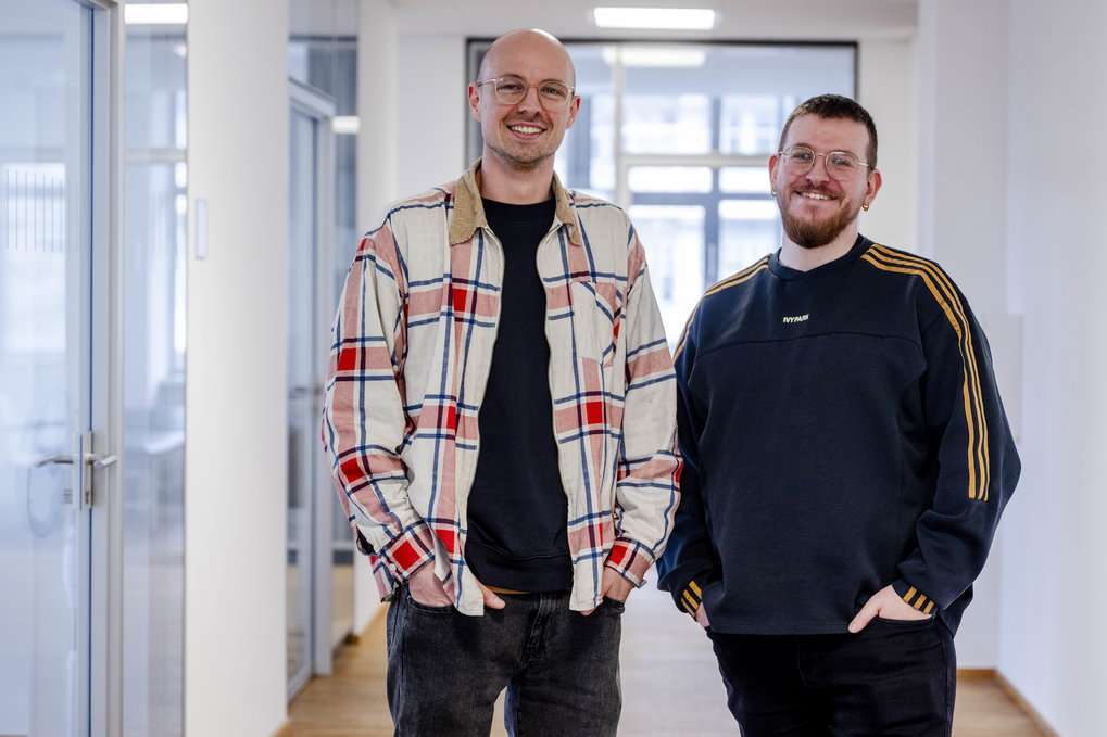 Two men in casual clothing are standing in a modern office hallway.